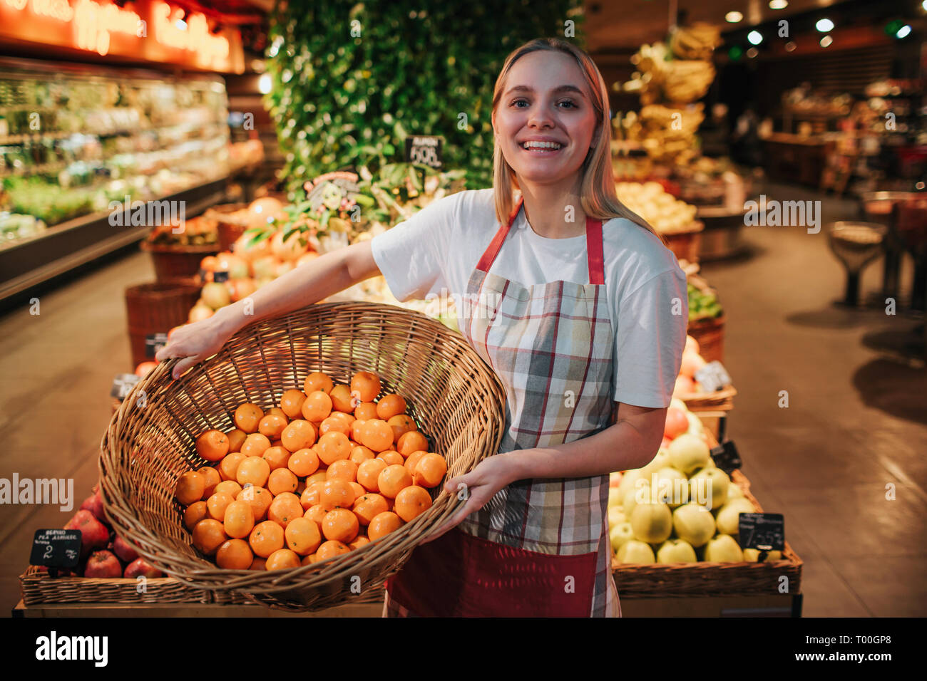 Young woman stand at fruit boxes in grocery store. She hold basket with oranges and pose on camera. Wears apron. Positive worker smile. She looks Stock Photo