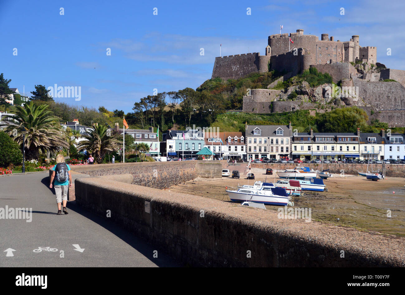 Woman Hiker Walking next to the Sea Wall at Gorey Harbour and Mount Orguell Castle on the Coastal Path, Island of Jersey, Channel Isles, UK. Stock Photo