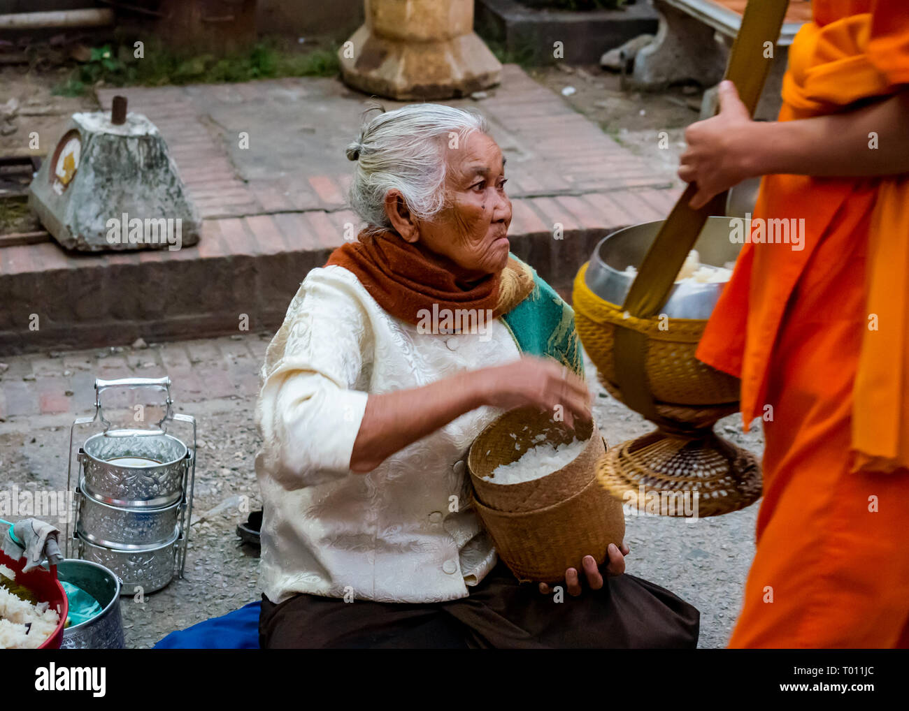 Old woman handing rice to Buddhist monk in orange robe in morning alms giving ceremony, Luang Prabang, Laos Stock Photo