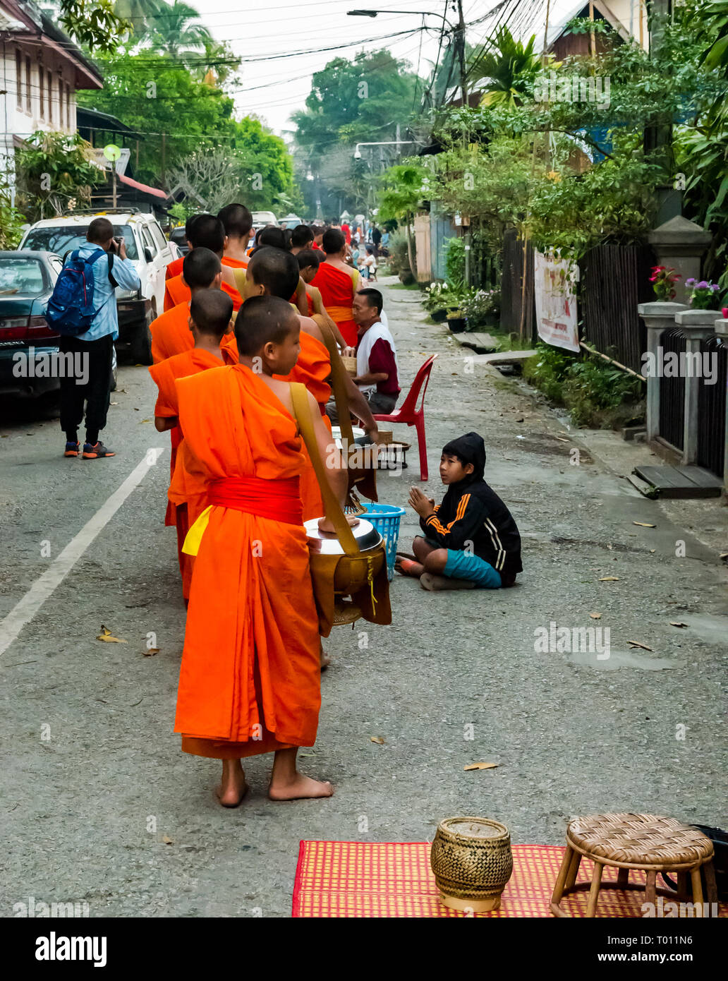 Buddhist monks in orange robes queue for morning alms giving ceremony with local poor boy collecting extra rice for his village, Luang Prabang, Laos Stock Photo