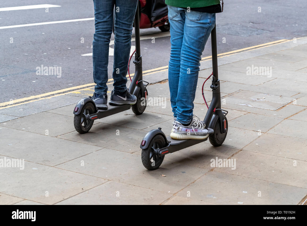Two adults ride electric scooters on the pavement, High Street Kensington, London Stock Photo