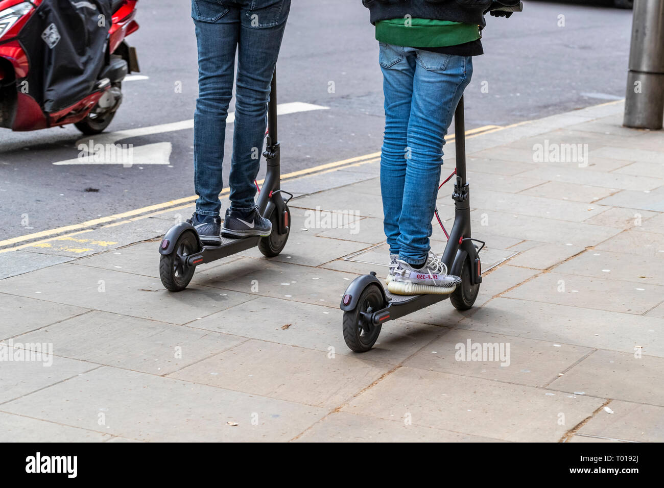 Two adults ride electric scooters on the pavement, High Street
