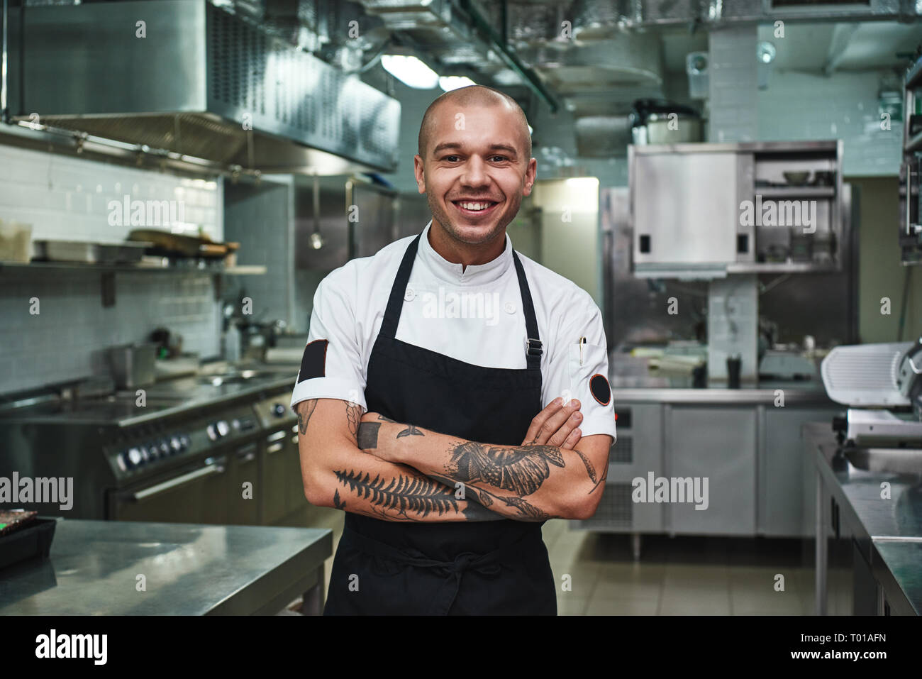 https://c8.alamy.com/comp/T01AFN/i-love-my-work-cheerful-young-chef-in-apron-keeping-tattooed-arms-crossed-and-smiling-while-standing-in-a-restaurant-kitchen-cooking-concept-T01AFN.jpg