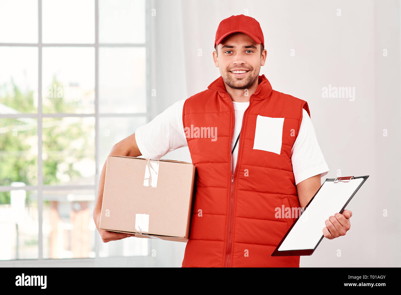 Cheerful postman wearing red postal uniform is delivering parcel to a client.  He holds carton box, folder and looking at the camera with a smile.  Friendly worker, high quality delivery service. Indoors.