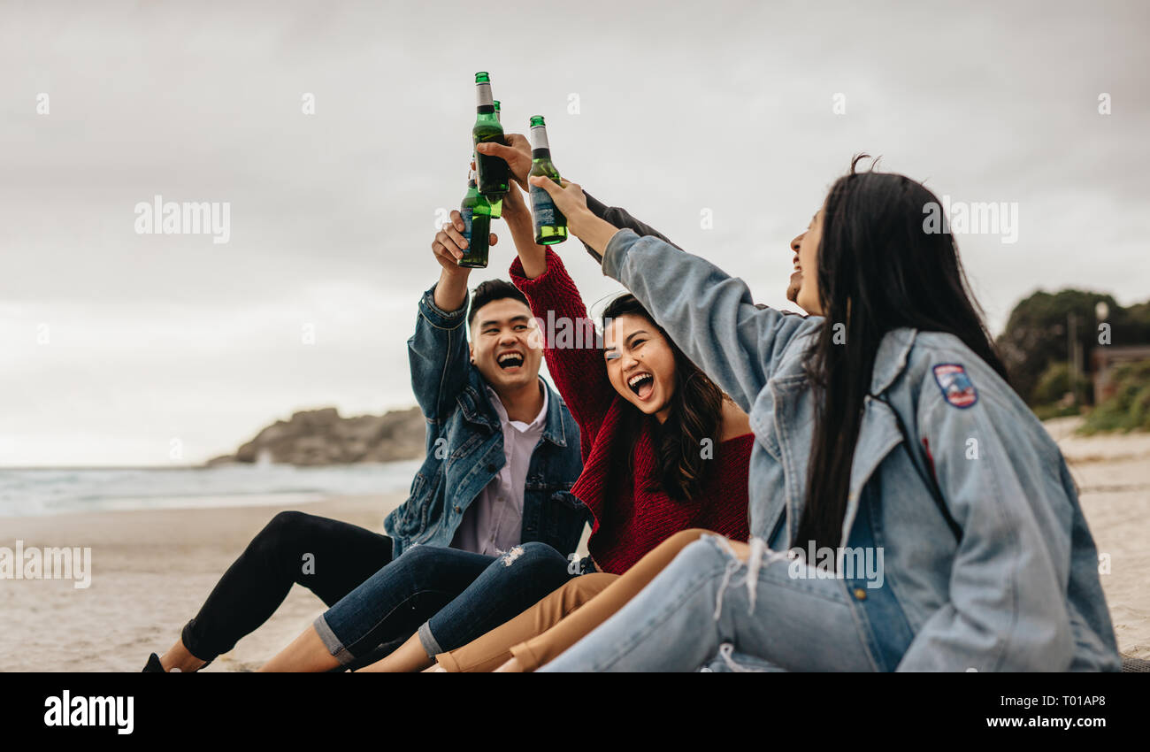 Group of four asian friends drinking beer on the sea shore. Young people on the beach having a party with drinks. Stock Photo