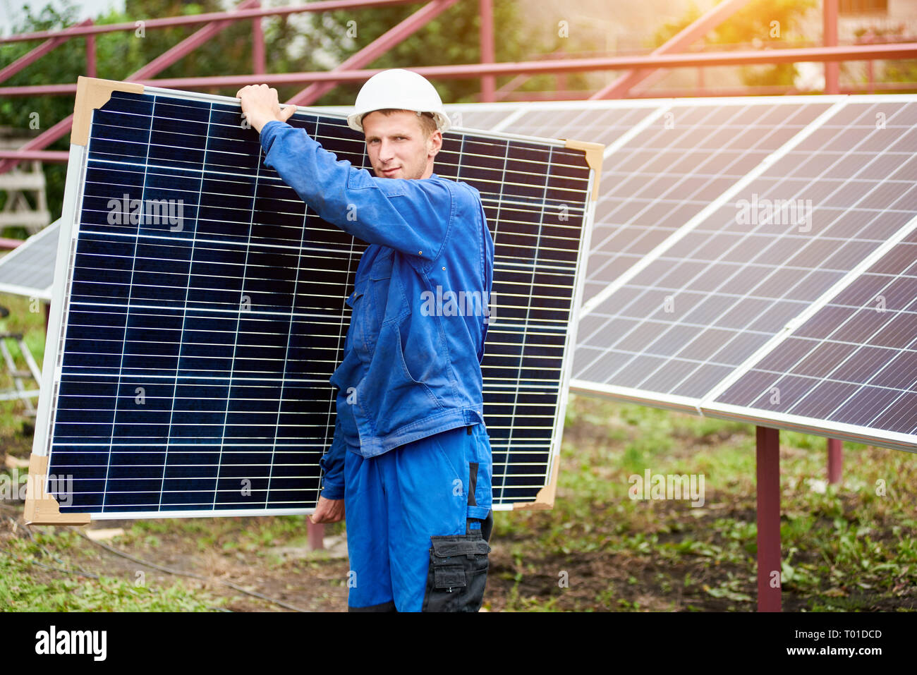 Portrait of worker in blue uniform and protective helmet carrying big shiny solar photo voltaic panel to almost finished exterior metal platform on su Stock Photo