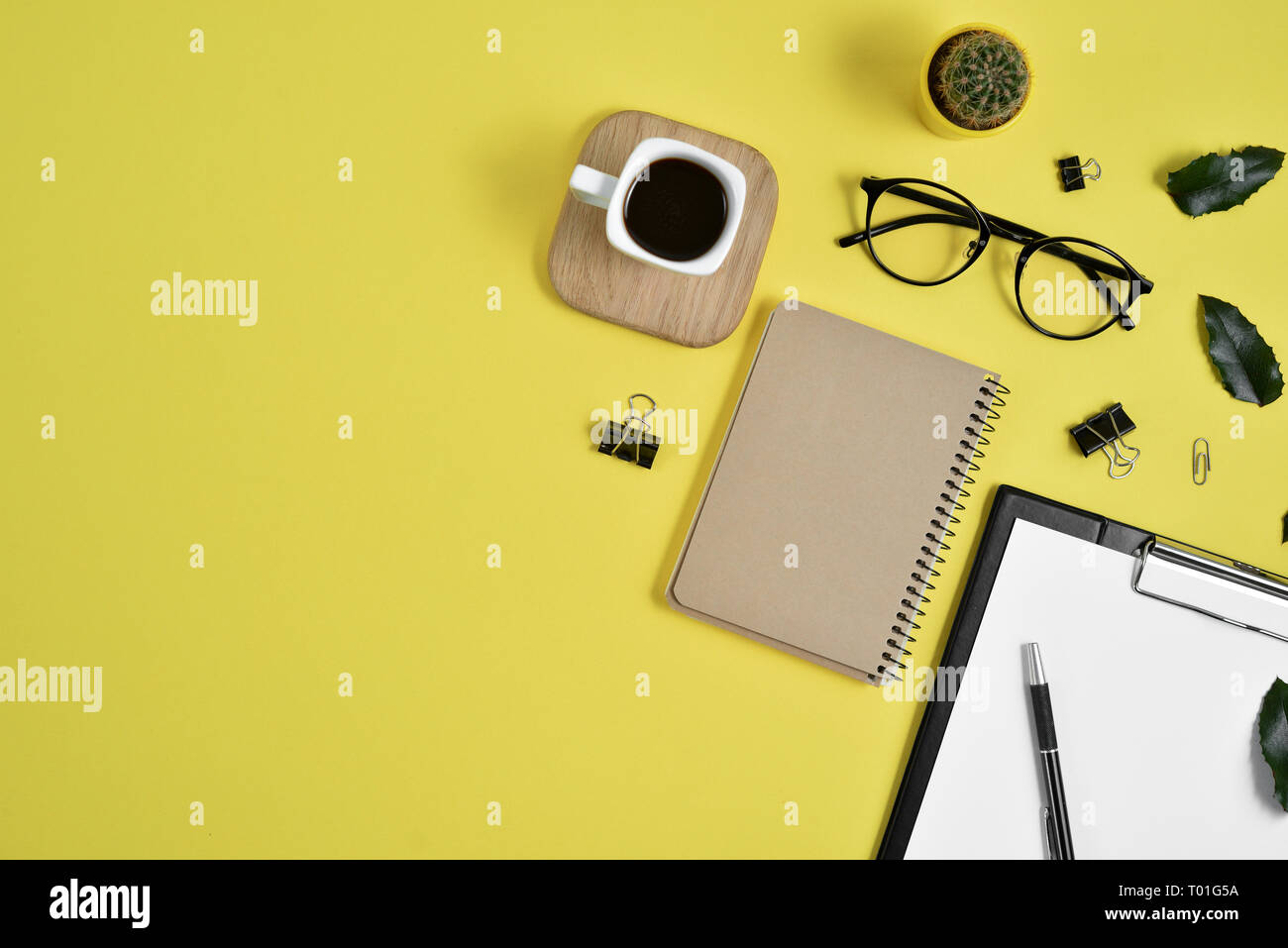 Office desk workspace with blank clip board, office supplies, pen, cactus, green leaf, coffee cup on a wooden stand and eye glasses on yellow backgrou Stock Photo