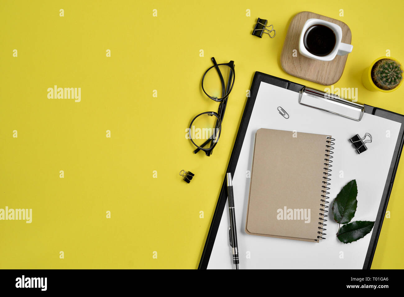 Office desk workspace with blank clip board, office supplies, pen, cactus, green leaf, coffee cup on a wooden stand and eye glasses on yellow backgrou Stock Photo