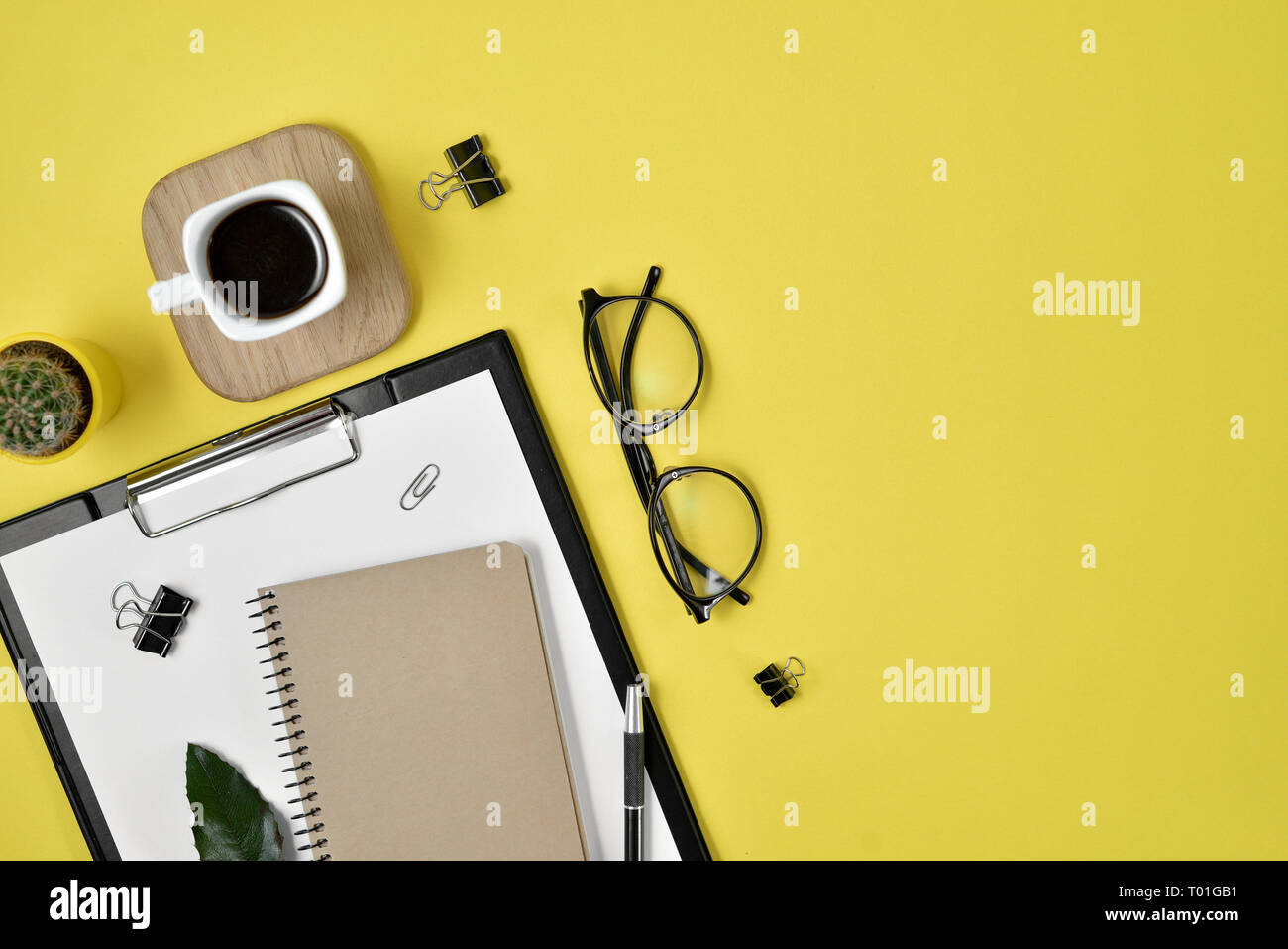 Office desk workspace with blank clip board, office supplies, pen, cactus, green leaf, coffee cup on a wooden stand and eye glasses on yellow backgrou Stock Photo