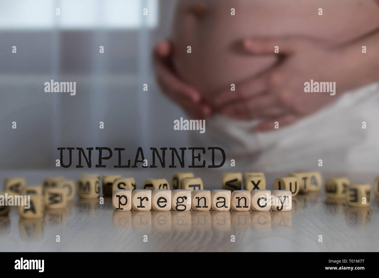 Words UNPLANNED PREGNANCY composed of wooden letters. Pregnant woman in the background Stock Photo