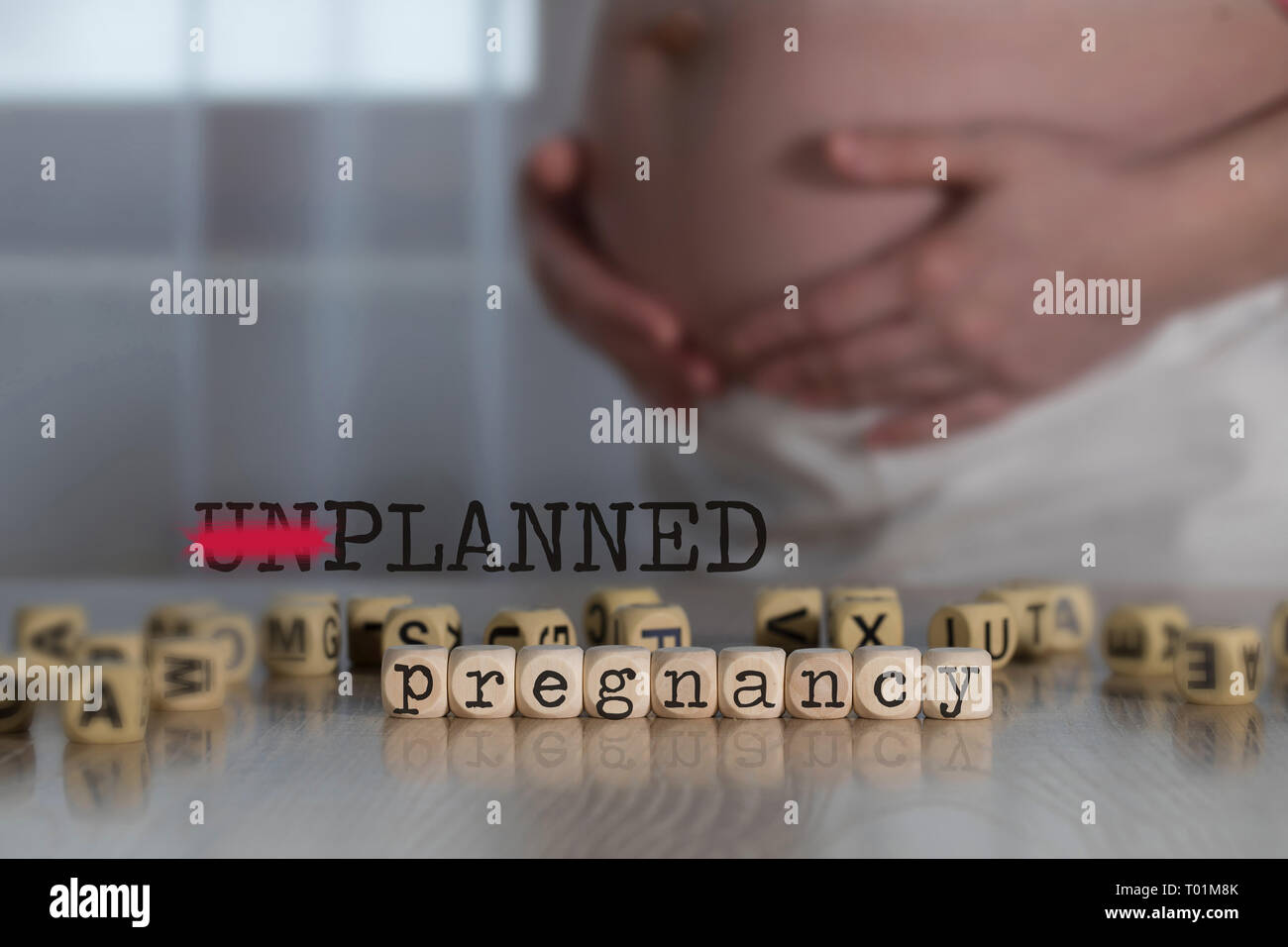 Words UNPLANNED PREGNANCY composed of wooden letters. Pregnant woman in the background Stock Photo