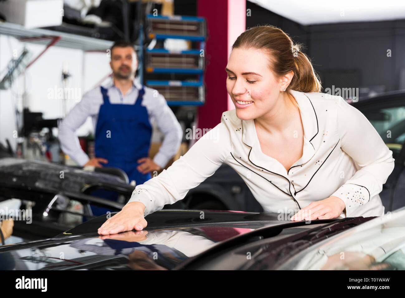 Happy young woman touching surface of repainted car body in auto repair shop Stock Photo