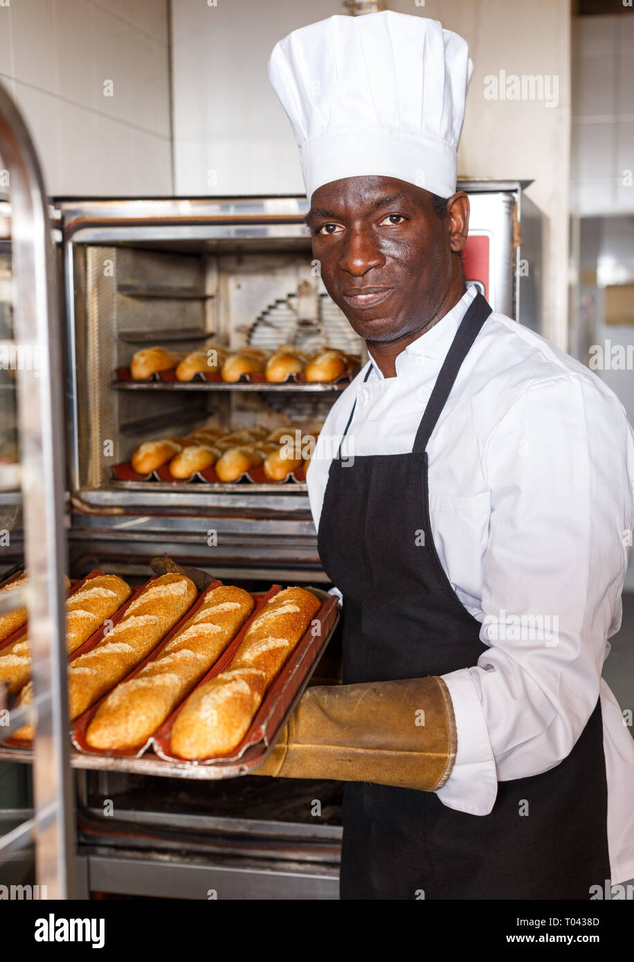 Experienced baker working in small bakery, taking out bread from industrial oven and putting on rack Stock Photo