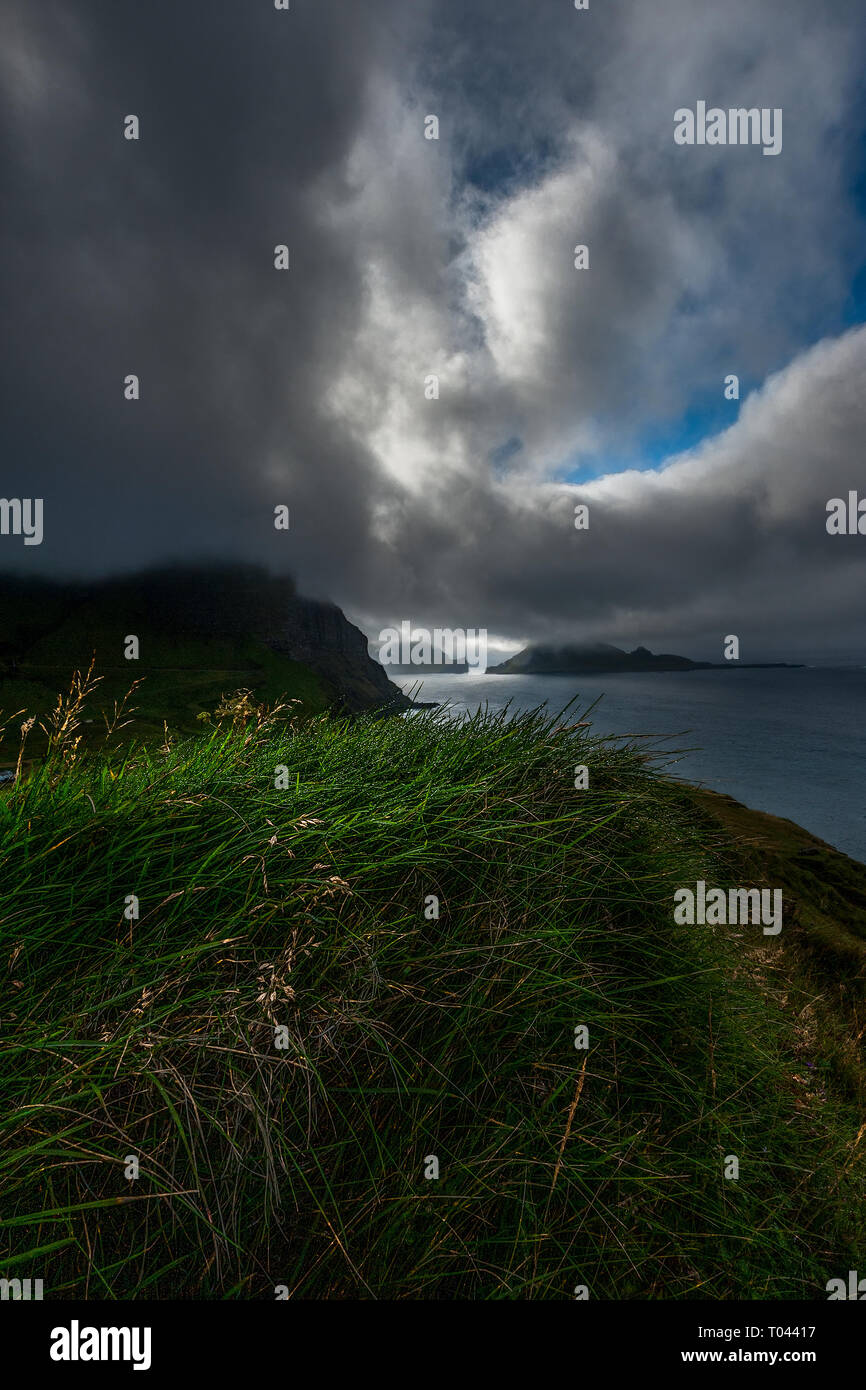 View from over the top of a green grassy hill towards the ocean and islets under a overcast blue sky with clouds, Faroe Islands. Stock Photo
