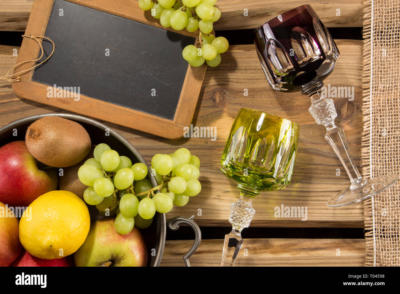 Traditional rummers/wine glasses on rustic wood planks and arranged fruits in a bowl, chalkboard and jute cloth. For wine menu or restaurant. Stock Photo