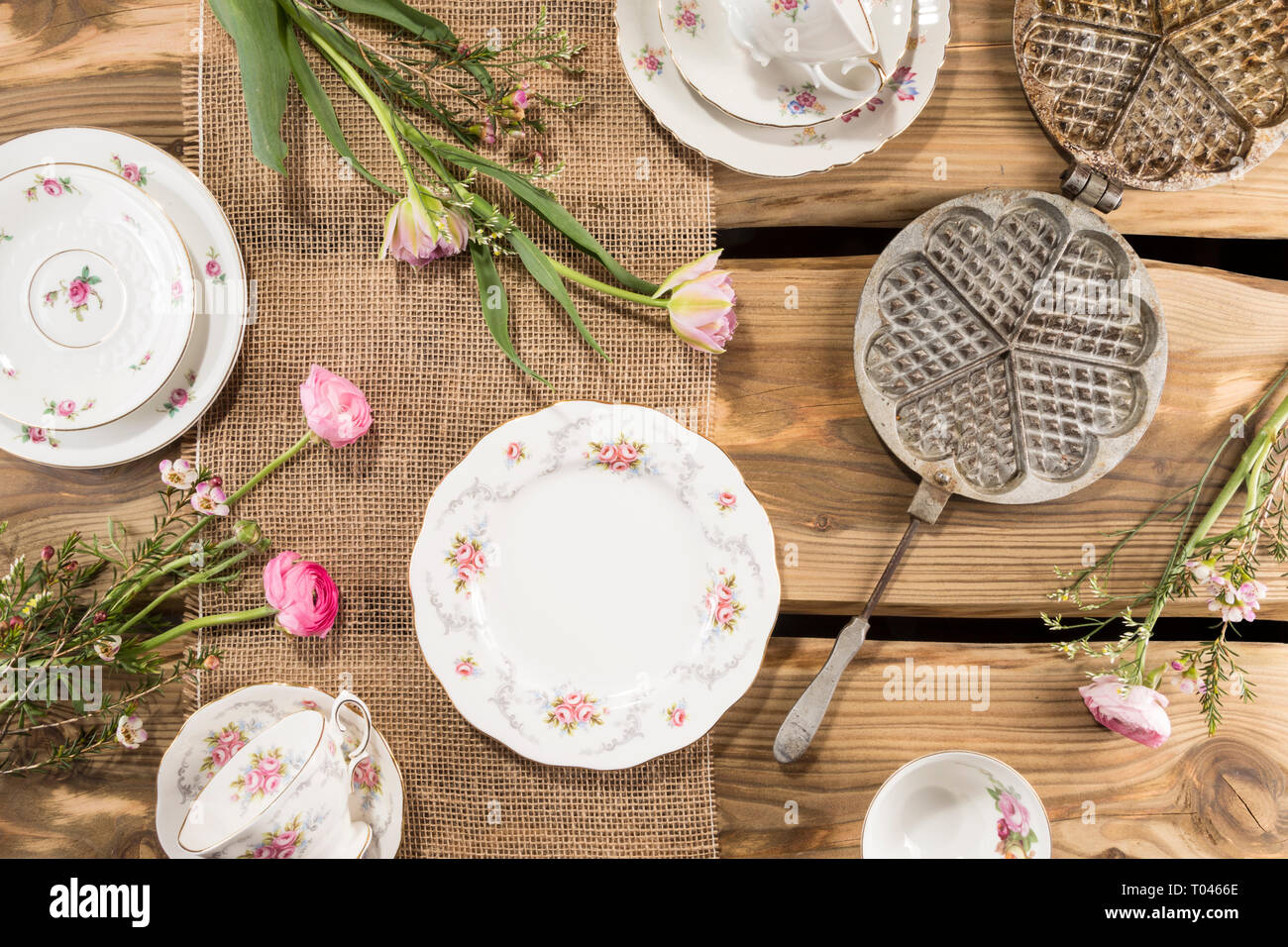 Old romantic porcelain, pink flowers and ancient waffle iron arranged on rustic wood planks. Shot from above, flat lay. Stock Photo