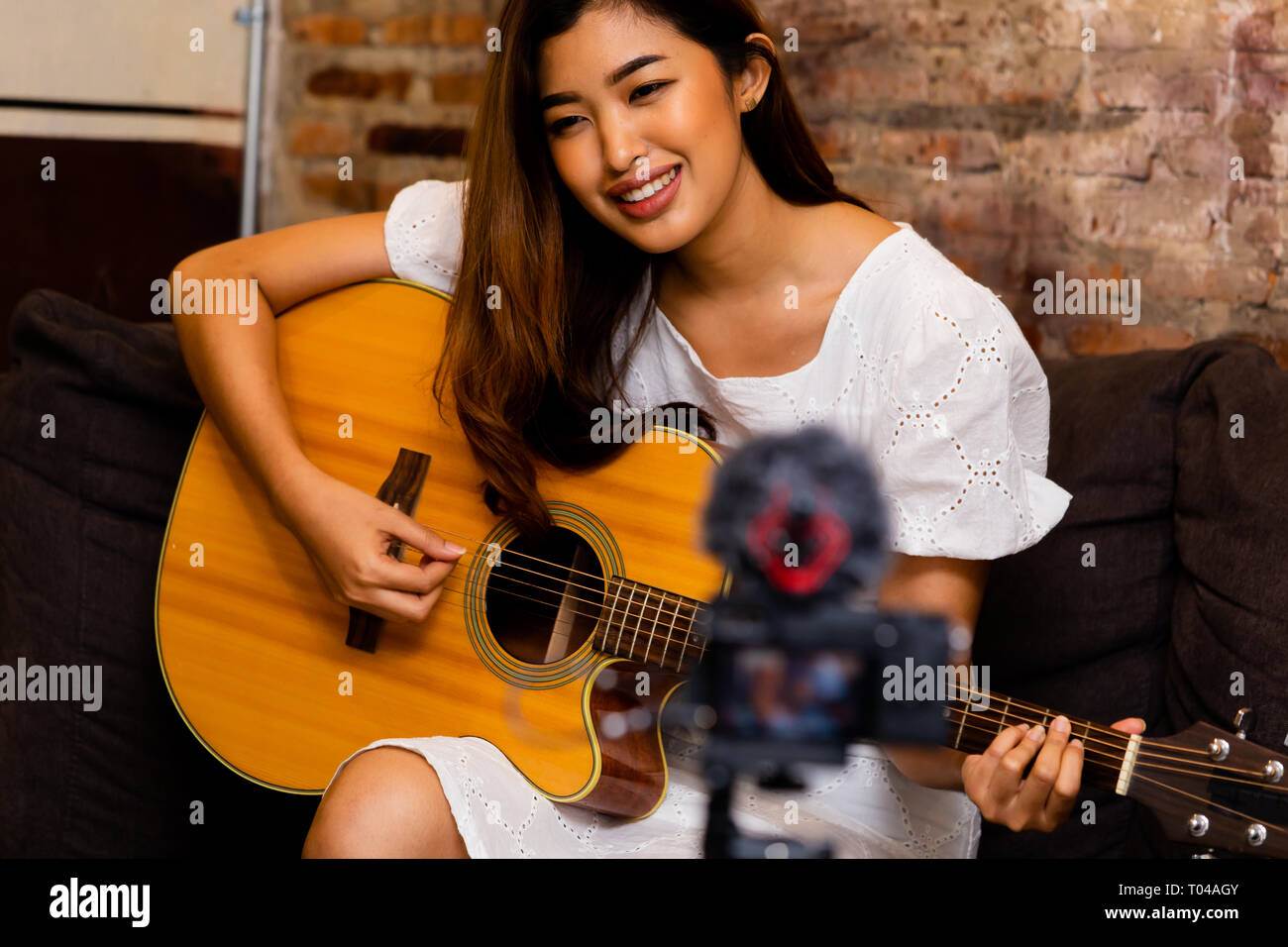 Lovely Asian female smiling and playing cover song while sitting on couch in front of video camera Stock Photo