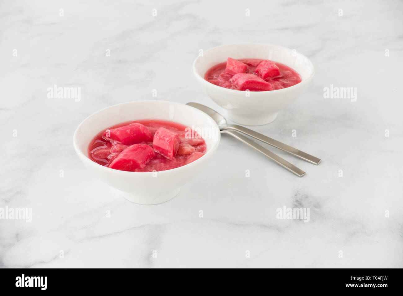 Homemade rhubarb compote, dessert, made from pink forced rhubarb, in two white porcelain bowls on white marble background. Stock Photo