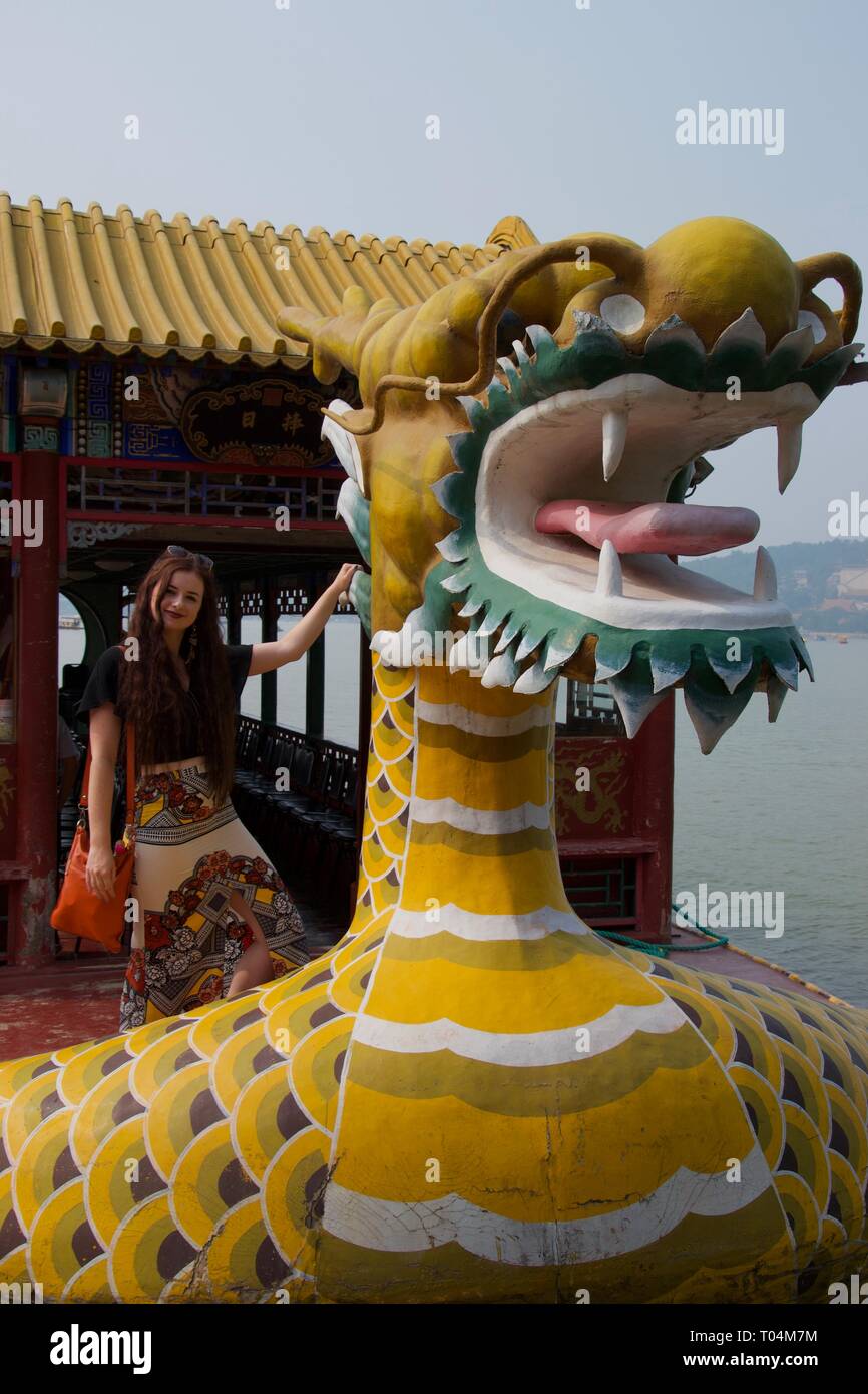 Western fashionable female tourist poses with yellow dragon carving on Chinese dragon boat. Dragon has open mouth and tongue stuck out Stock Photo