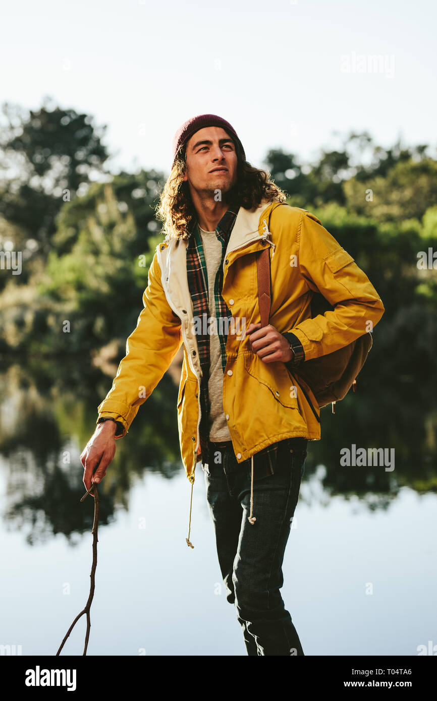Man exploring nature walking in a forest beside a lake. Young tourist carrying a backpack walking outdoors holding a stick. Stock Photo