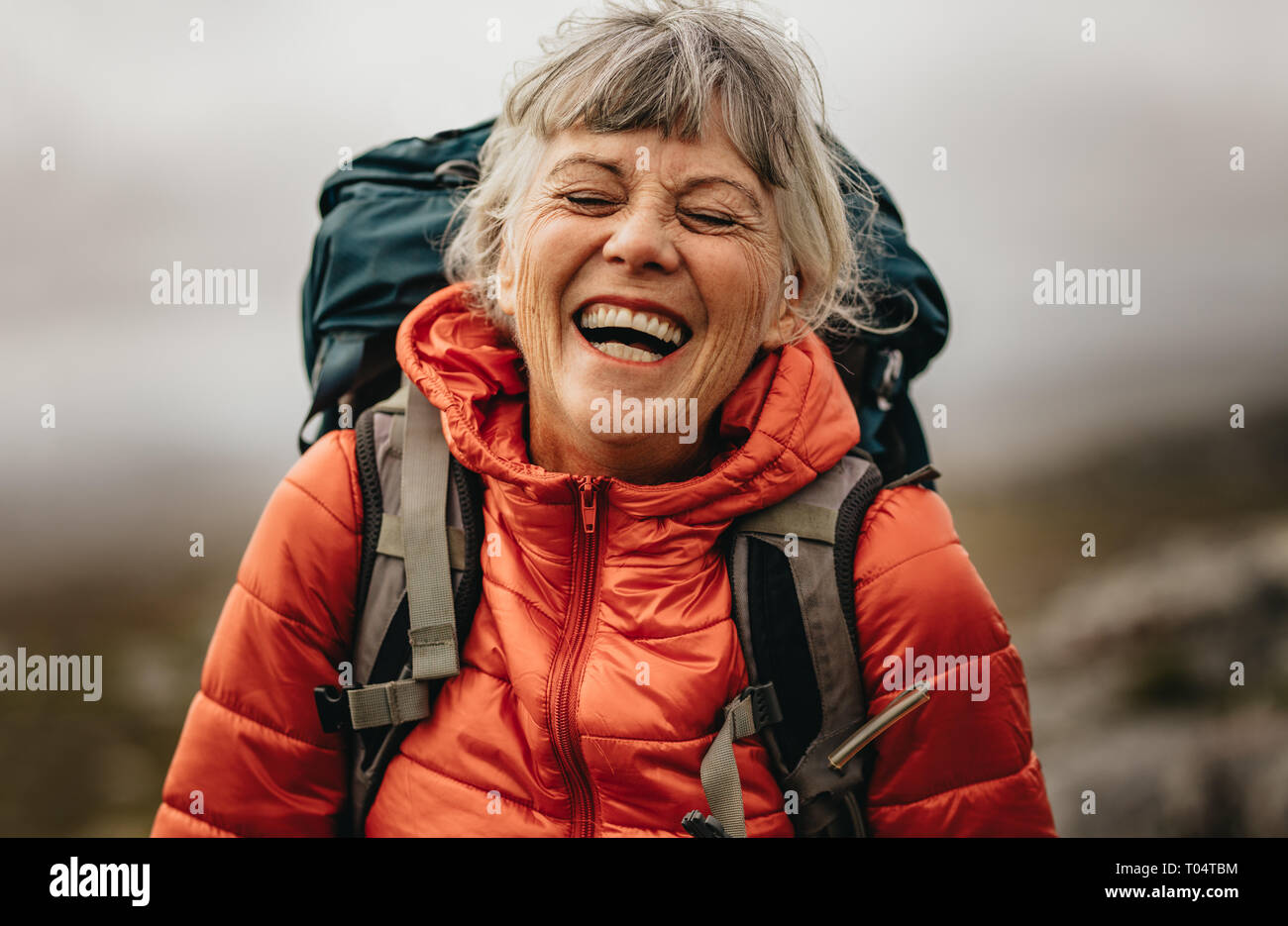 Close up of a senior woman wearing jacket and backpack laughing with eyes closed. Portrait of a woman hiker laughing during her trekking. Stock Photo