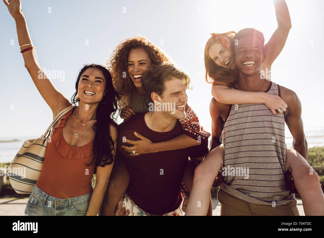 Two young men carrying female friends on their back and walking outdoors. Group of young friends having fun outdoors on a sunny day. Stock Photo
