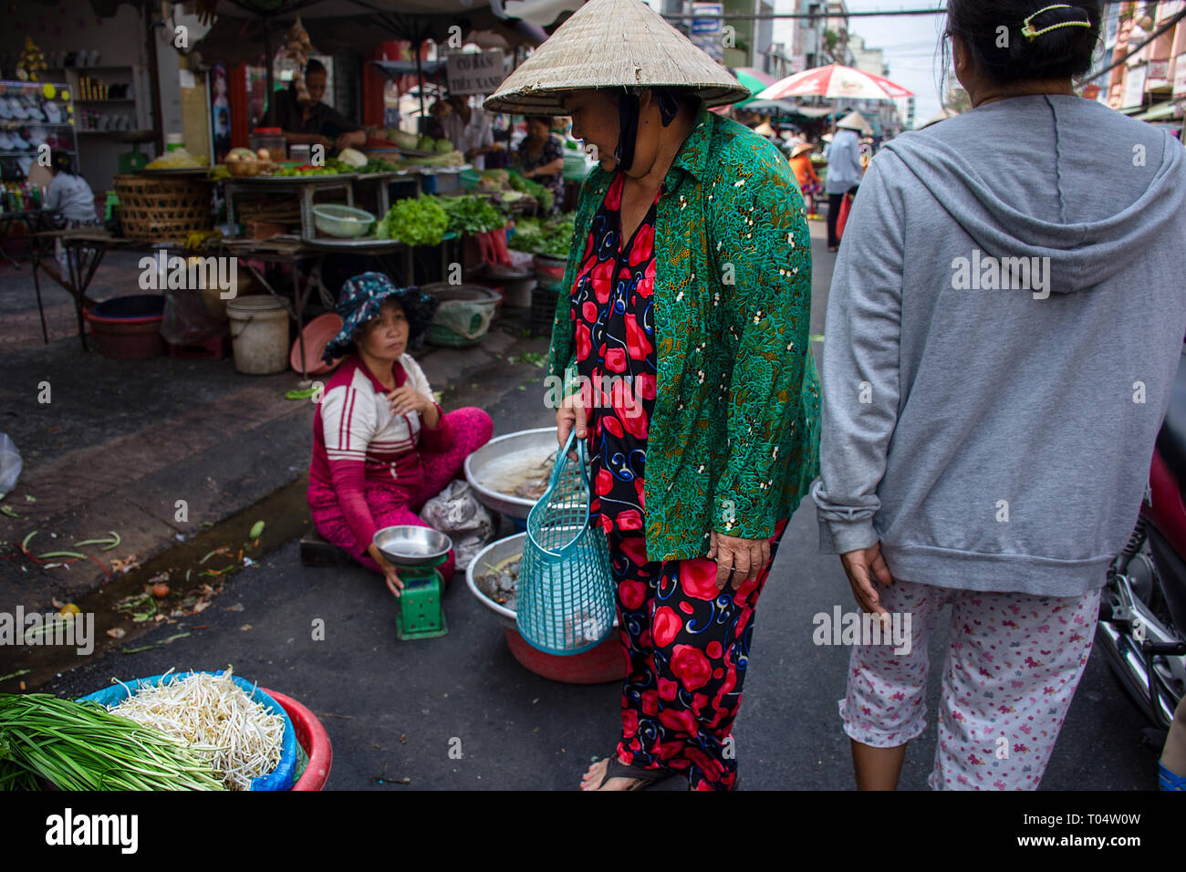 Elderly Asian woman in bright outfit with Chinese hat shopping for vegetables at a street market, Ho Chi Minh City, Vietnam. Stock Photo