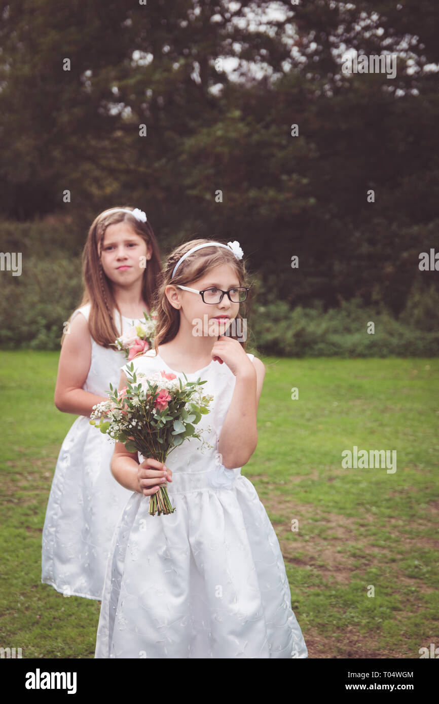 Sweet child and young teen girl bridesmaids wearing short white dresses, holding hand-tied bouquets with foliage in a park waiting Stock Photo