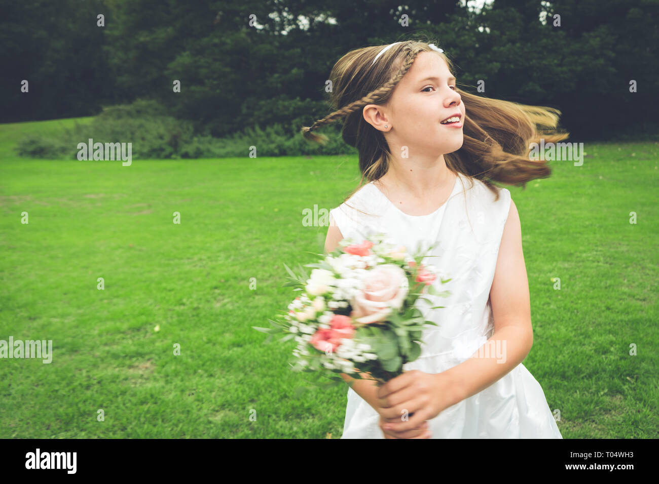 Young girl bridesmaid dancing and spinning in a park, wearing a white dress and holding a rustic posy Stock Photo
