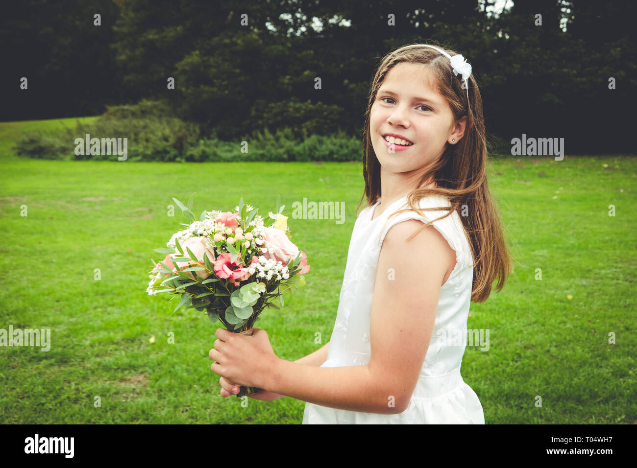 Sweet young girl tween or teen bridesmaid in a white dress with rustic posy bouquet in a park Stock Photo