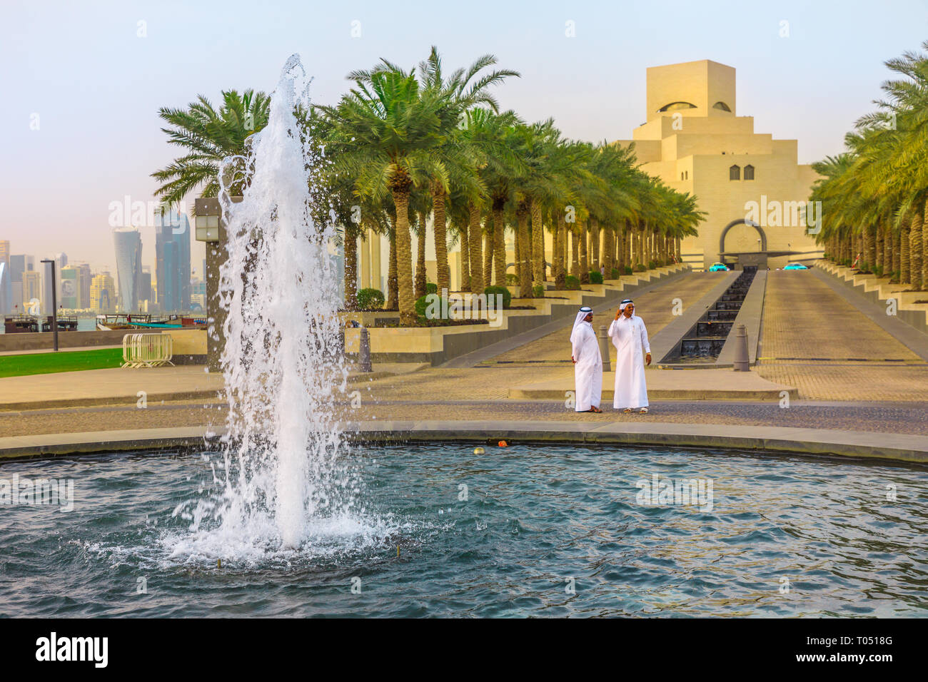 Doha, Qatar - February 16, 2019: two Arabs standing near fountain of Museum of Islamic Art with skyscrapers of West Bay skyline on background. Middle Stock Photo