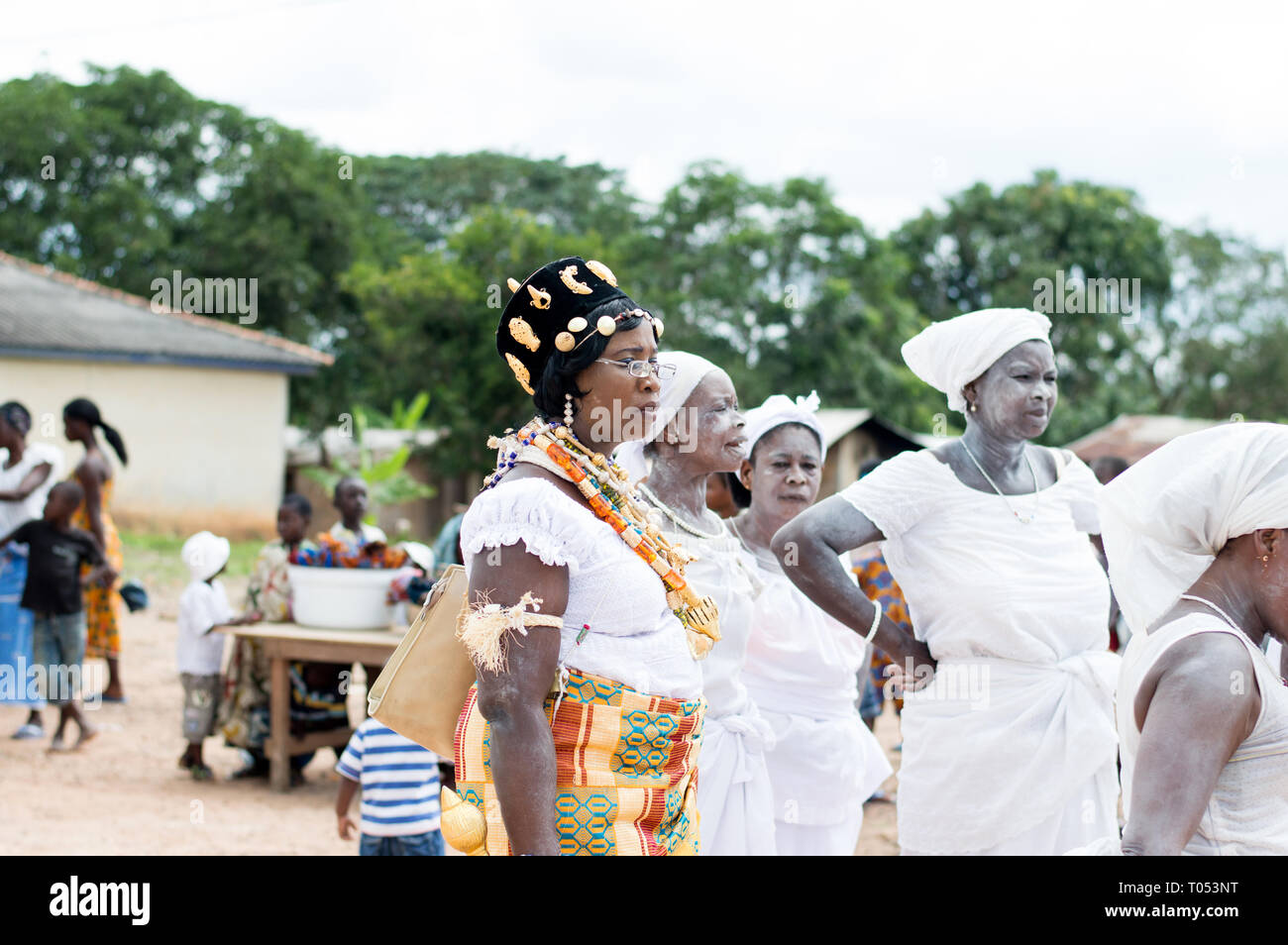 adzopé, ivory coast-august 31, 2016: young woman dressed in traditional dress, gold hat glasses neck collars standing near other women in white and lo Stock Photo