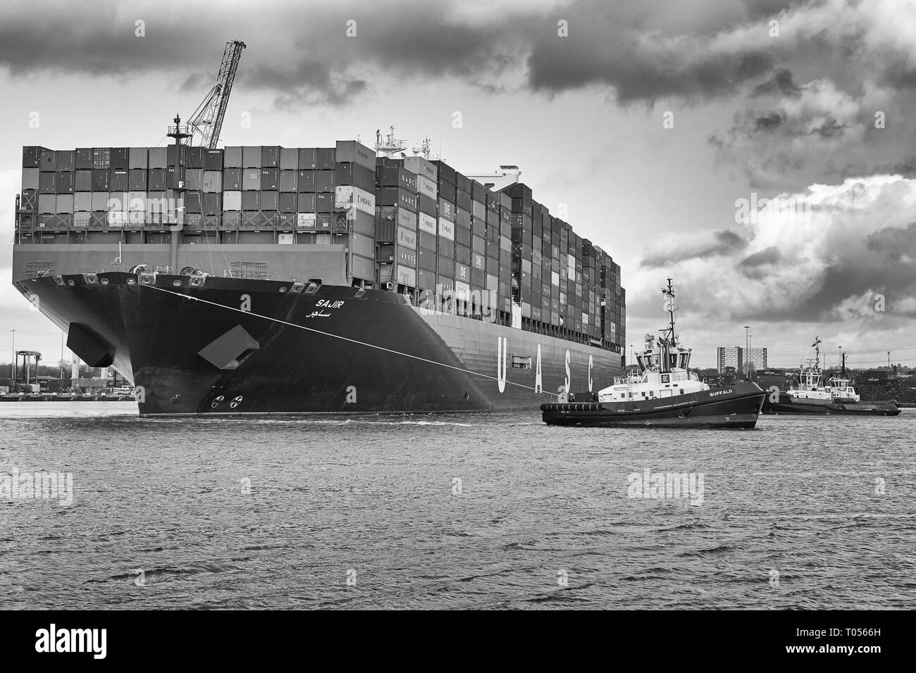 Black & White Image Of Three Tugs Assisting The Ultra-Large UASC Container Ship, SAJIR, Departing The Southampton Container Terminal, Hampshire, UK. Stock Photo