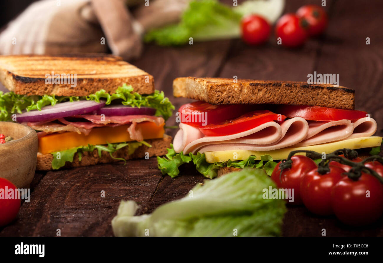 Fresh delicious toasts with cheese, tomato, onion, ham, prosciutto, cucumber and lettuce on wooden table and dark background Stock Photo