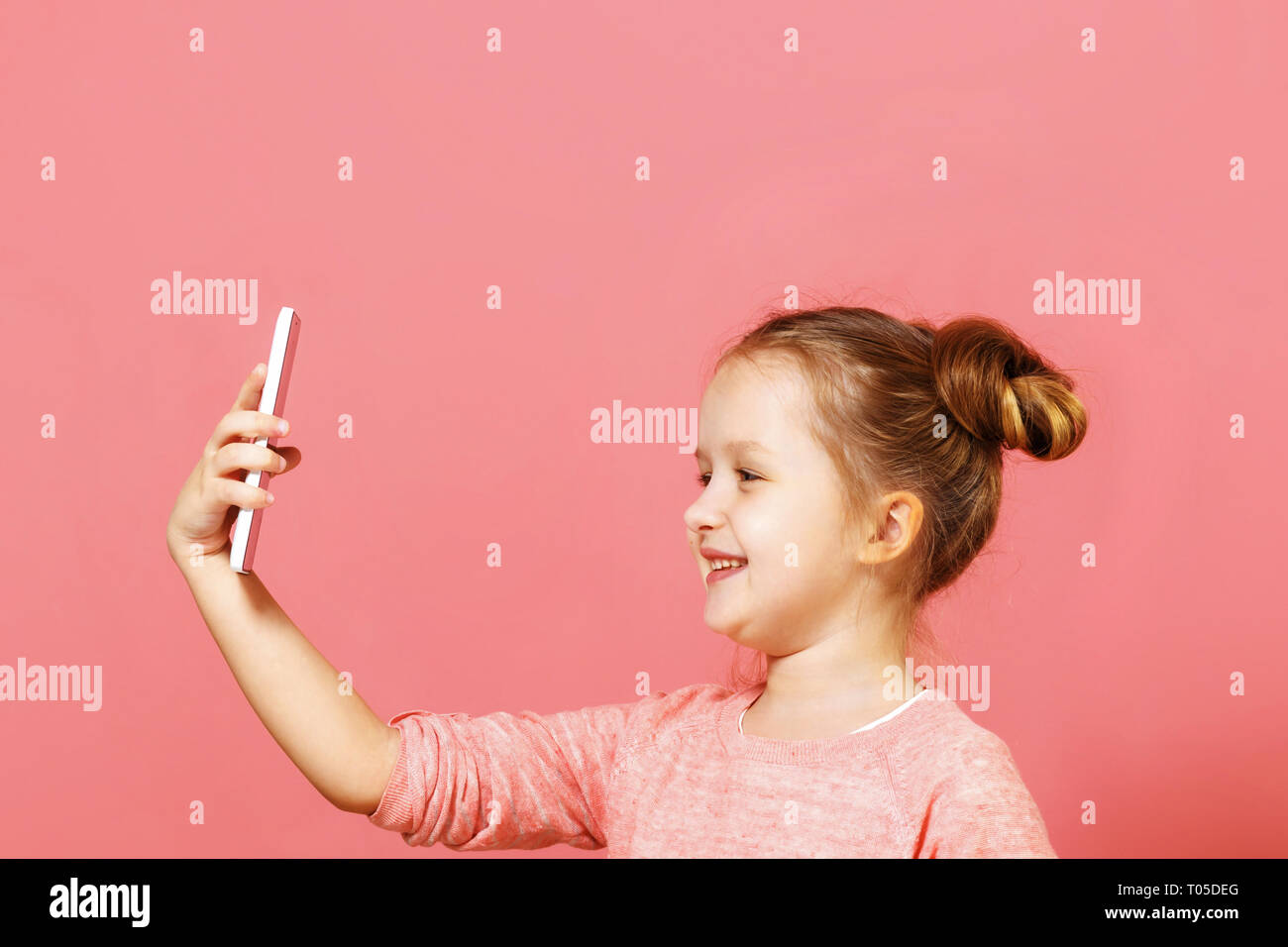 Closeup portrait of a cute little girl with buns of hair on a pink background. The child holds the phone and takes a selfie Stock Photo