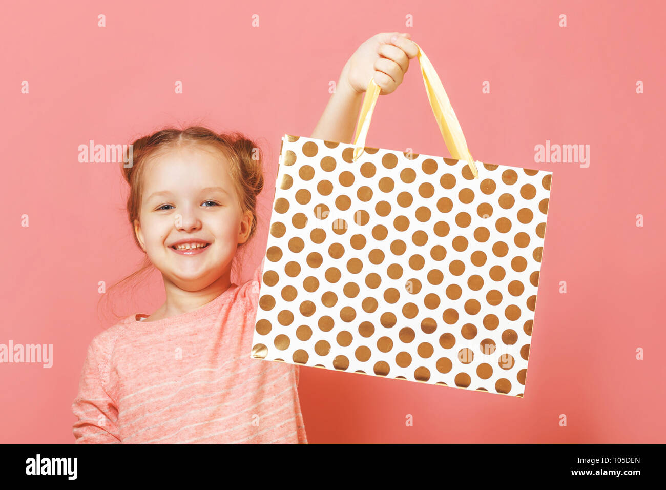 Closeup portrait of a cute little girl with hair buns over pink background. The child lifted the paper bag high, rejoicing at the shopping or gift. Stock Photo