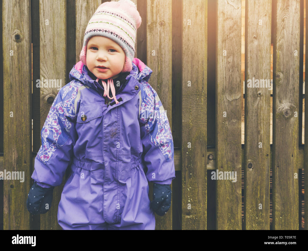 little girl wearing purple coverall standing by the wooden fence Stock Photo