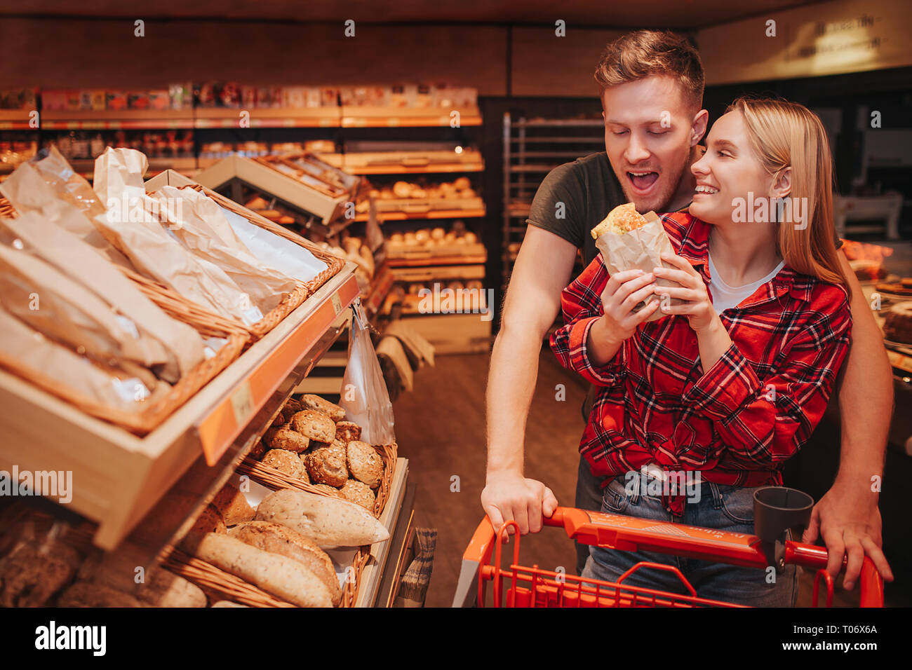 Young couple in grocery store. Funny man hold trolley with hands and want to bite roll. Woman hold it and smile. People stand at bread shelf. Stock Photo