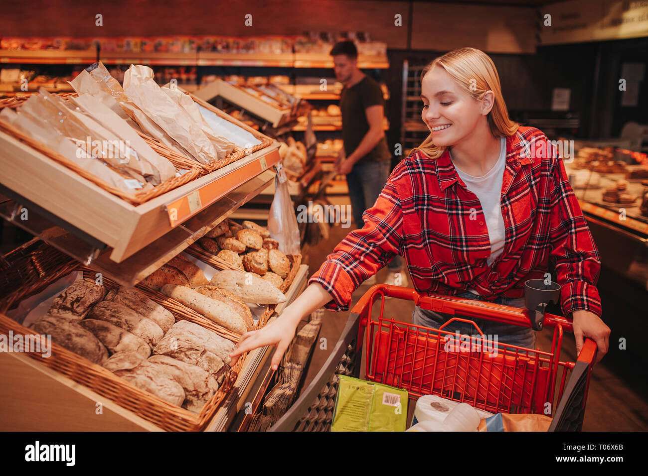 Young couple in grocery store. Woman reach to bread with hand and smile. Guy stand behind and pick food. Stock Photo