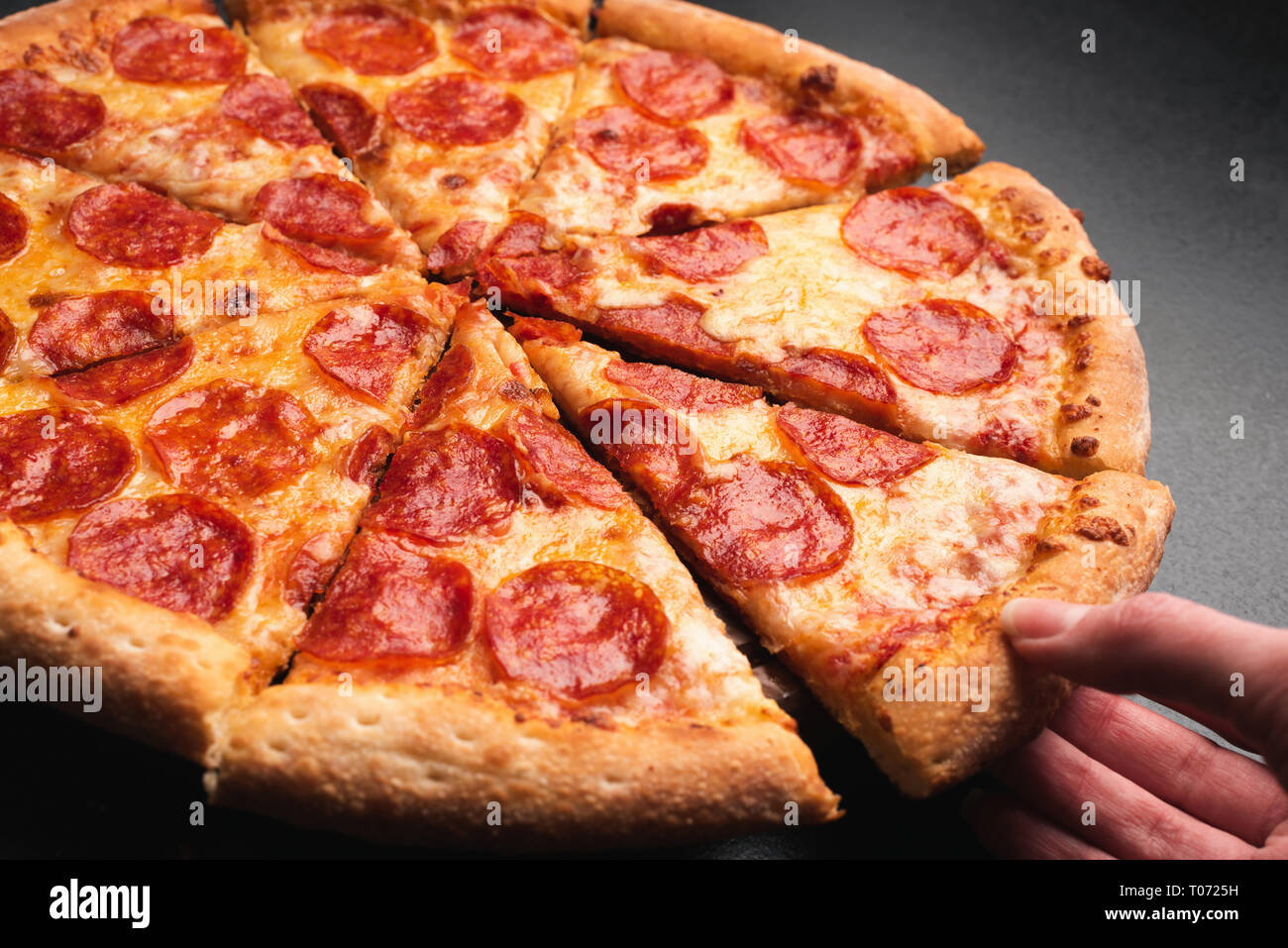 Taking slice of pepperoni pizza. Tasty pepperoni pizza on black background, closeup view. Hand taking pizza Stock Photo