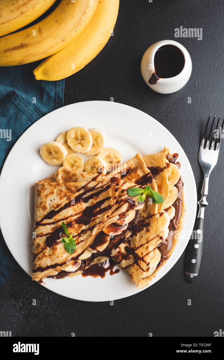 Crepes with chocolate spread and banana on white plate on black concrete background. Table top view Stock Photo