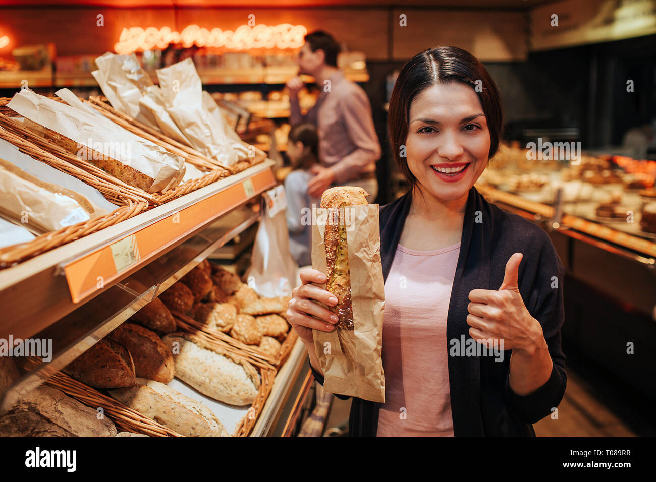 Young parents and daughter in grocery store. Positive cheerful woman hold bread in hands and show big thumb up. Fathr and daughter stand behind and Stock Photo