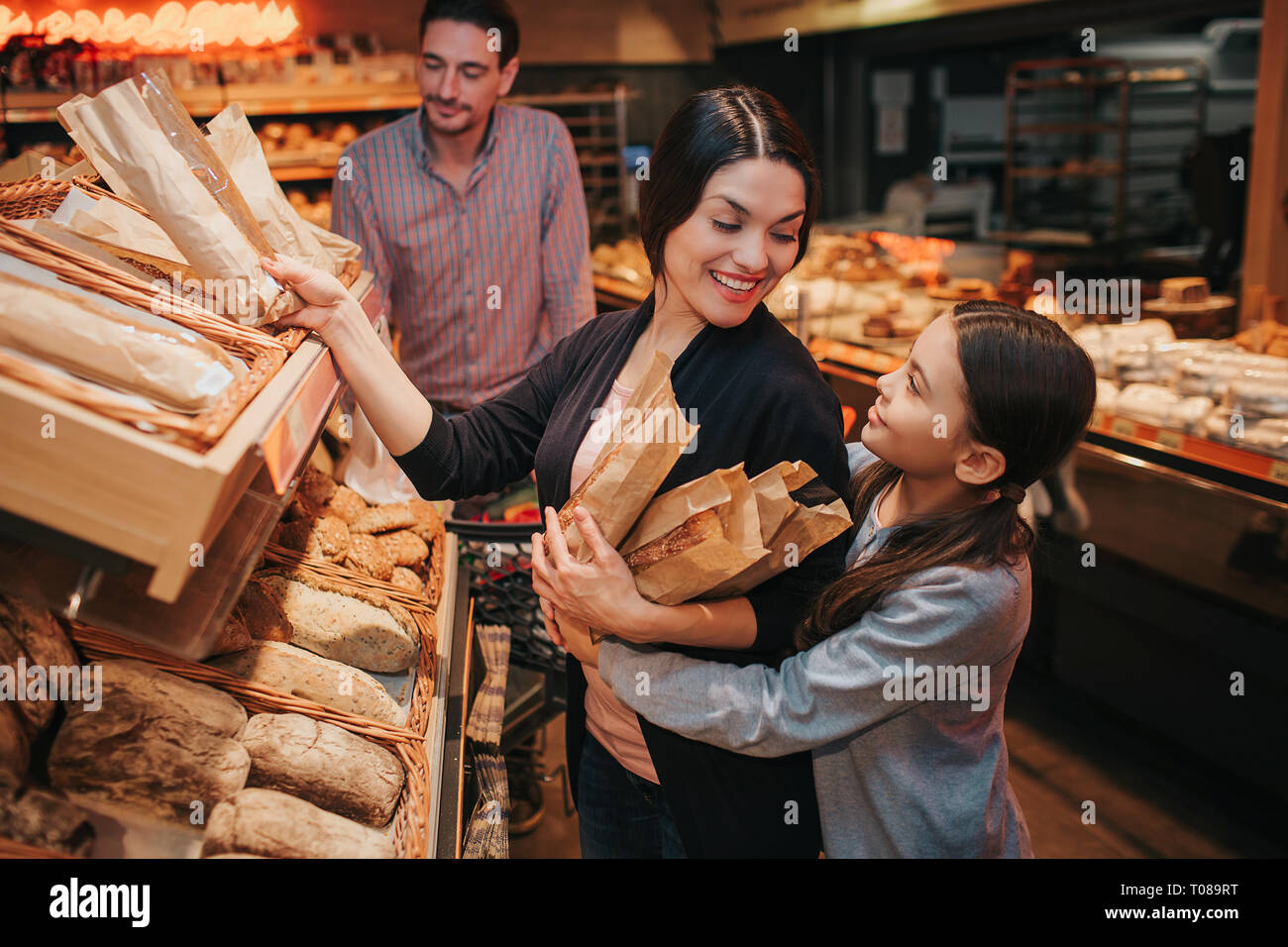 Young parents and daughter in grocery store. Cheerful mother and girl pick upb bread and rolls together. They smile to each other. Man stand behind Stock Photo