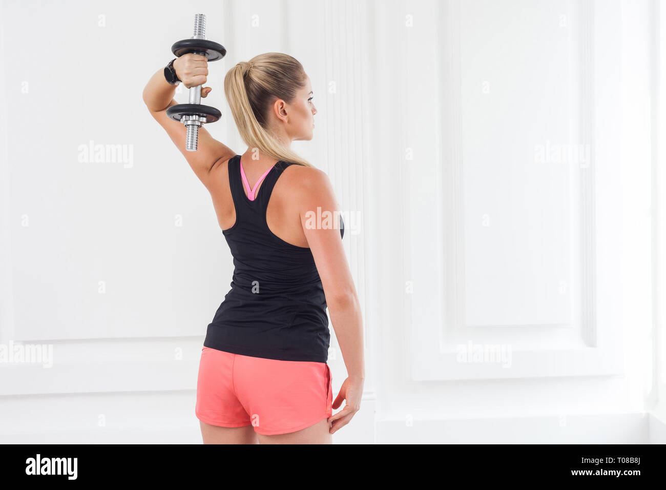 Striving for strong. Backside of young adult motivation athletic bodybuilder woman in pink shorts and black top standing, holding dumbells doing exerc Stock Photo