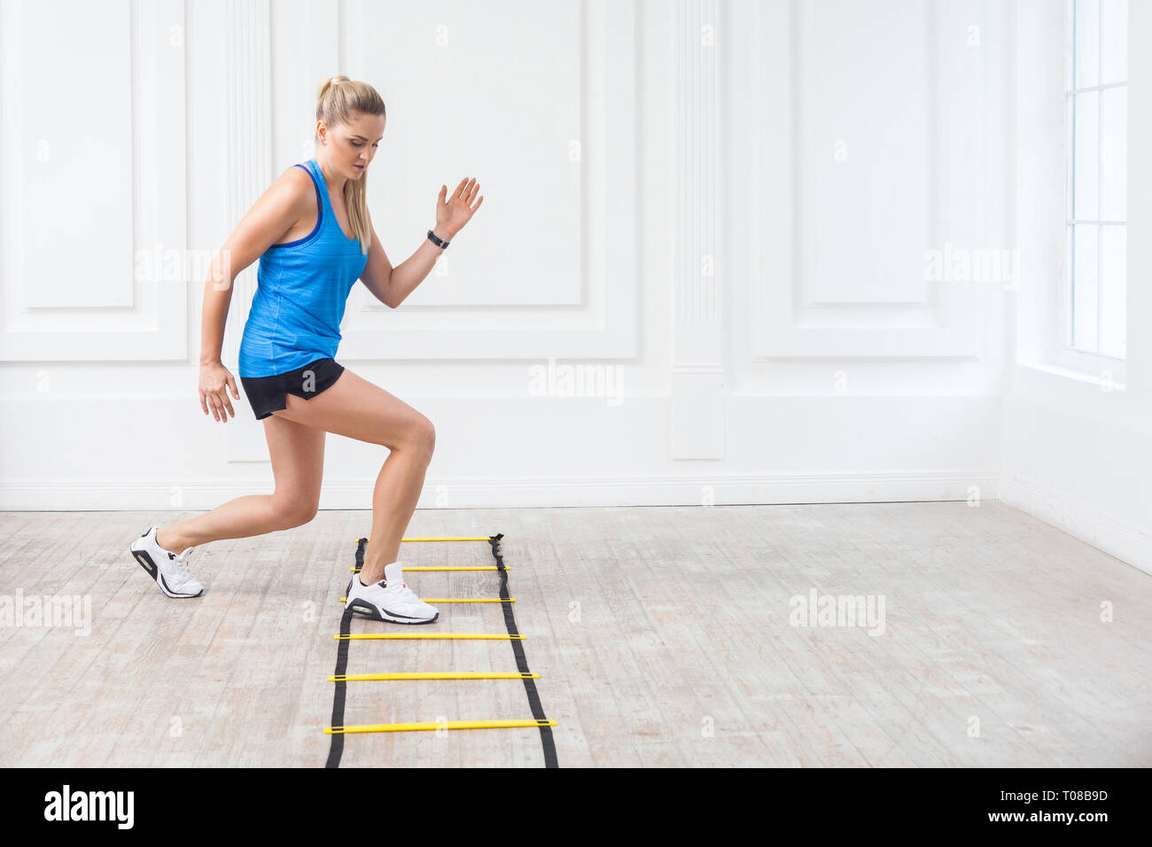 Full length of sporty beautiful young athletic blonde woman in black shorts and blue top are hard working and training on agility ladder drills in gym Stock Photo