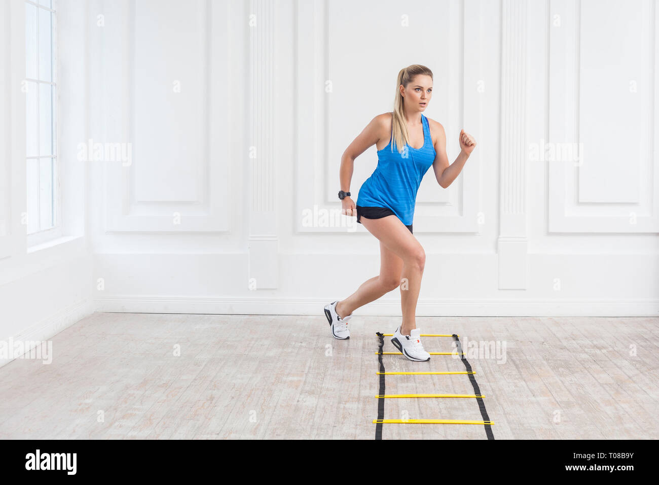 Full length of sporty beautiful young athletic blonde woman in black shorts and blue top are cardio workout with speed straps on floor and training on Stock Photo