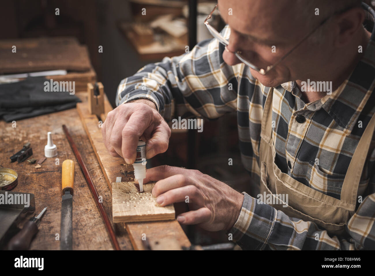 Mature artisan sitting at a rustic workbench while carefully applying glue on a small item Stock Photo