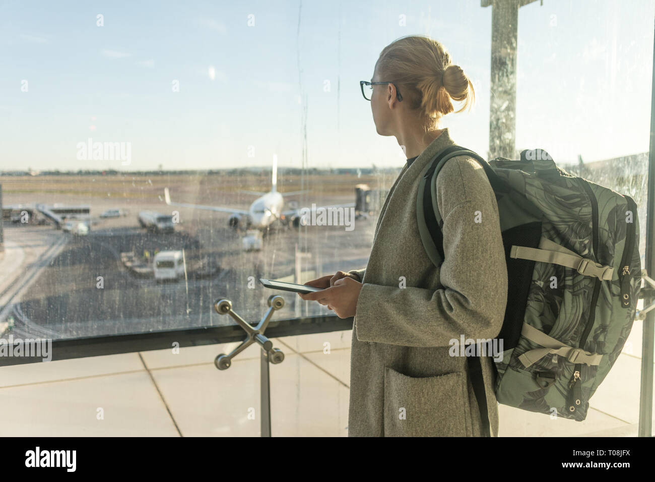 Casually dressed female traveler at airport, holding smart phone device, looking through the airport gate windows at planes on airport runway Stock Photo