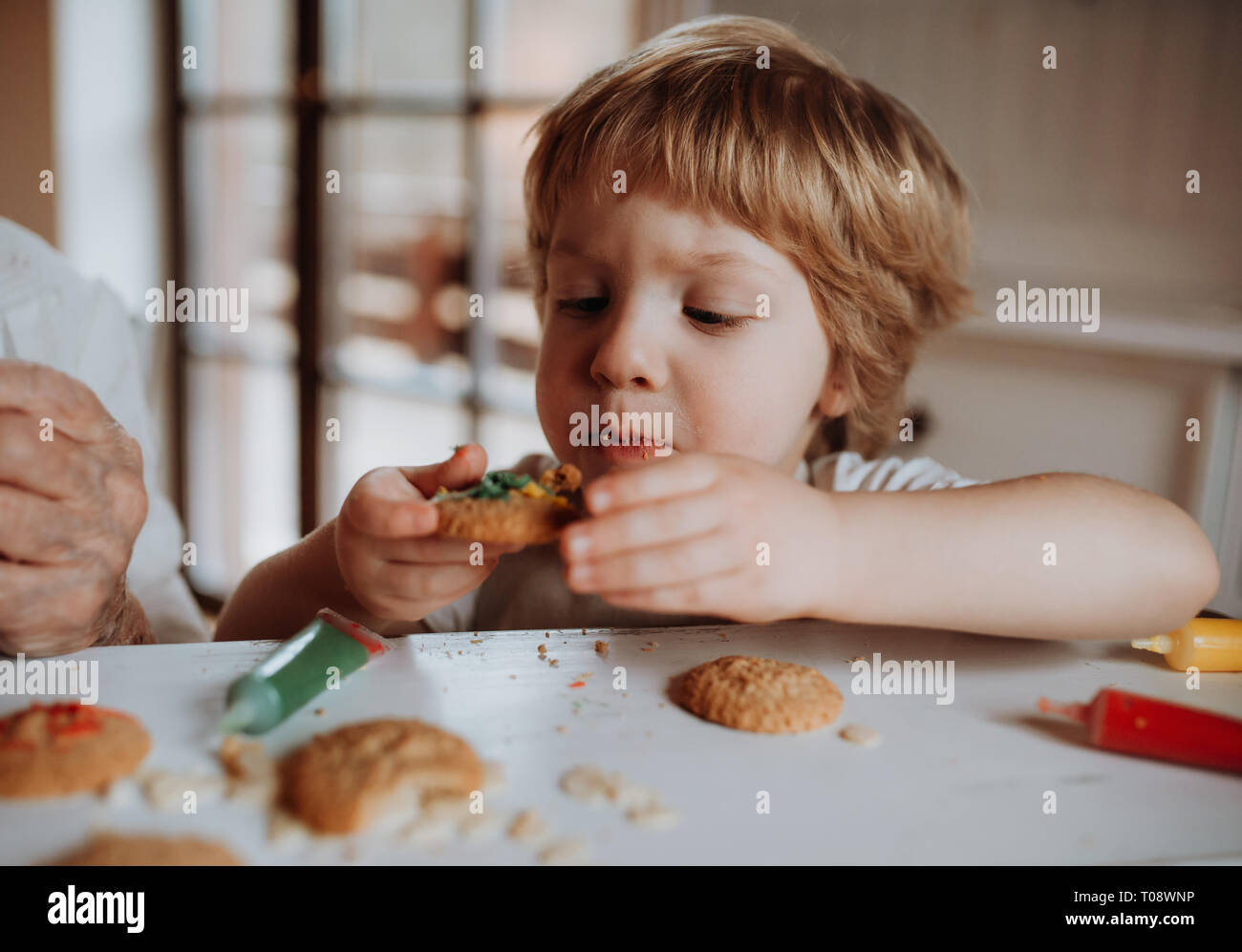 A small toddler boy sitting at the table, decorating and eating cakes at home. Stock Photo