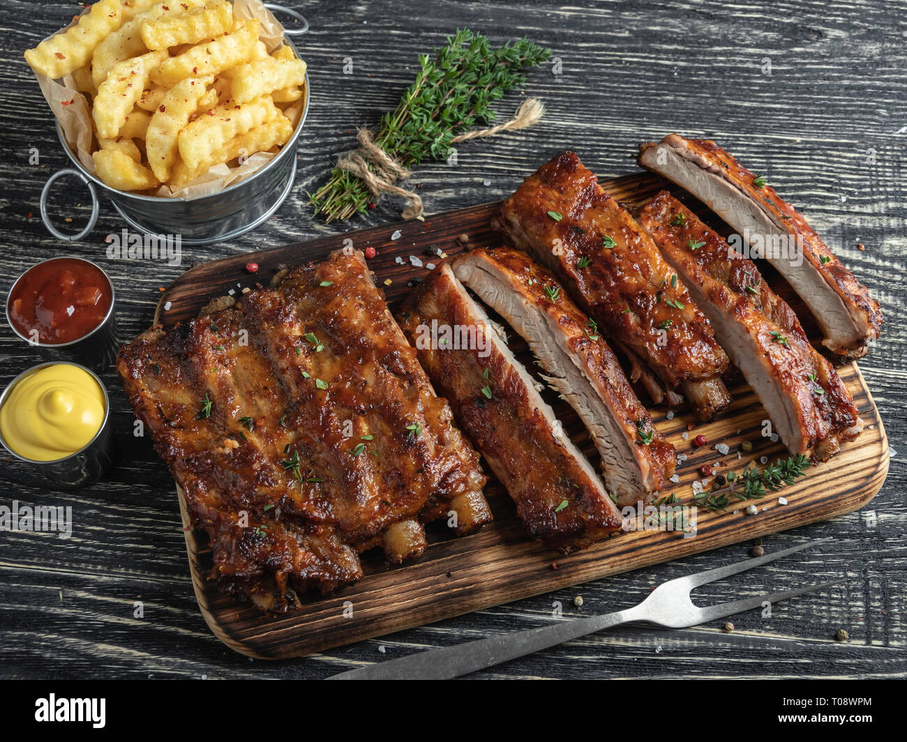 cutting grilled pork ribs with sauce on a board, french fries, spices, wooden background Stock Photo